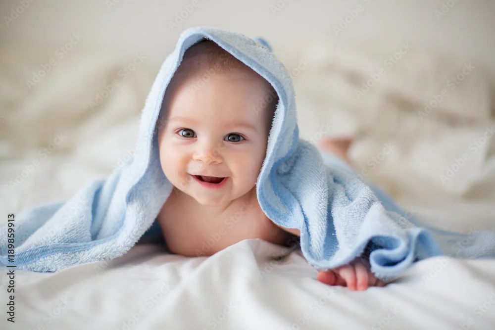 smiling baby boy with blue towel draped over his head, lying on a soft bedspread