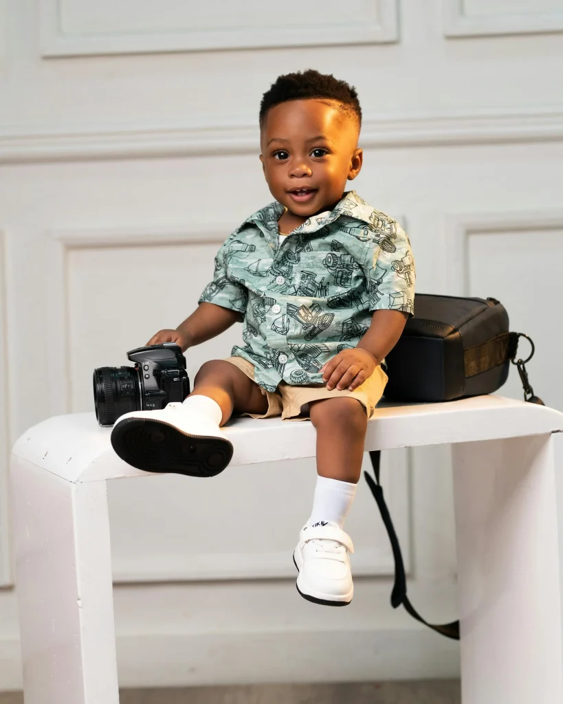 Cute baby boy sitting on a table, holding a camera, wearing a patterned shirt and shorts, with a cheerful expression.