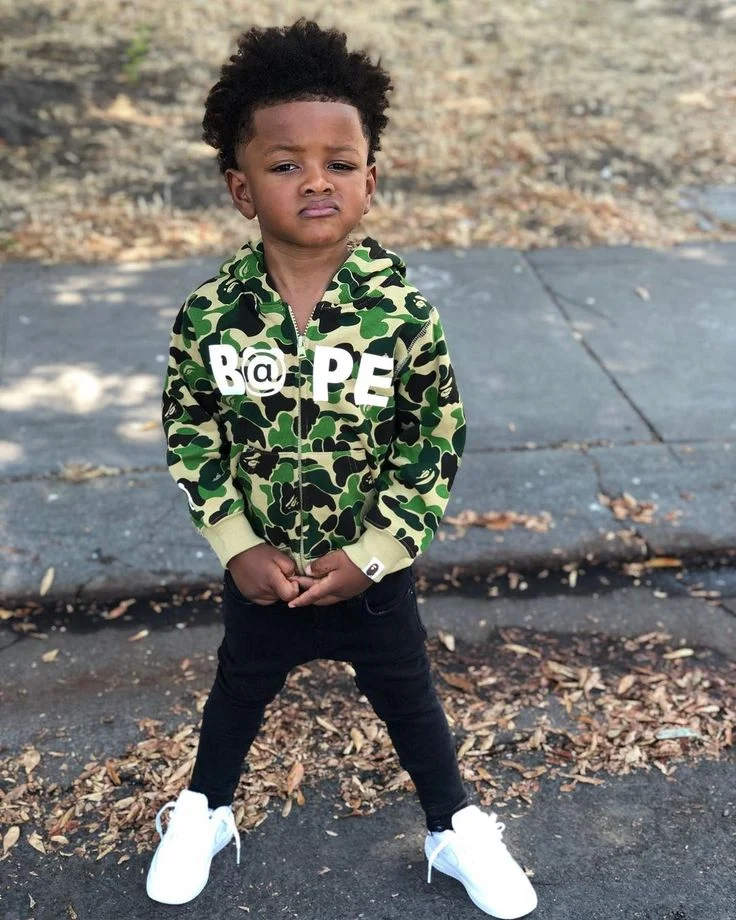 A young boy stands confidently in a camo hoodie and black pants, wearing white sneakers.
