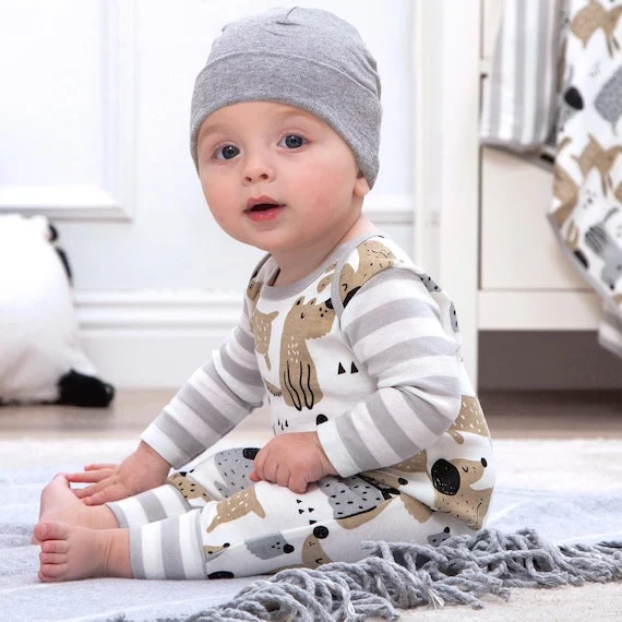 baby boy in a cozy animal-patterned romper and gray hat, sitting on a rug with a playful expression