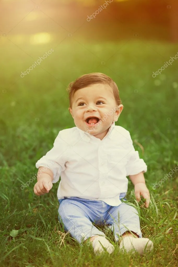happy baby boy in a white shirt sitting on grass, smiling with his tongue out in a sunlit setting