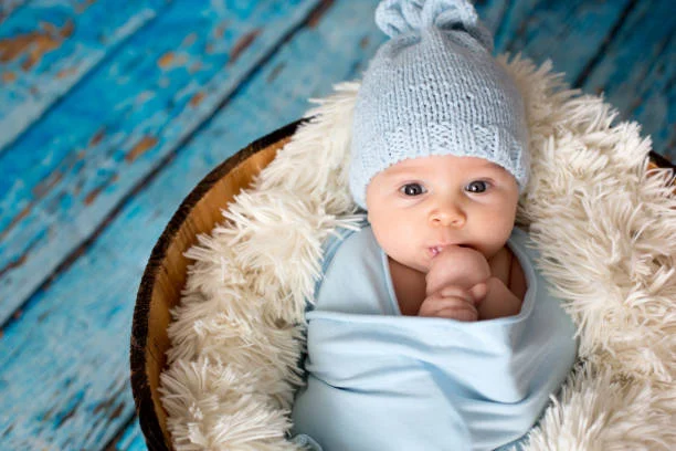 Newborn baby in a cozy basket, wearing a light blue wrap and knitted hat, with a soft background.