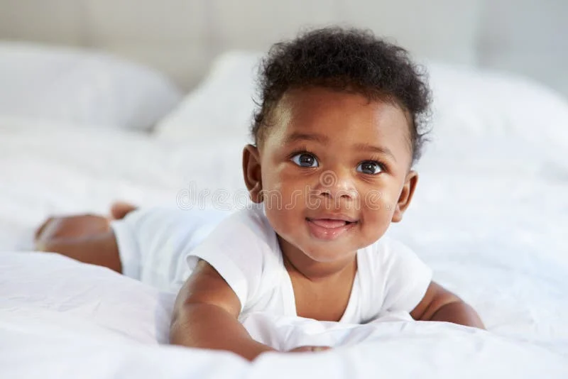 smiling baby with curly hair lying on a bed, surrounded by soft white bedding