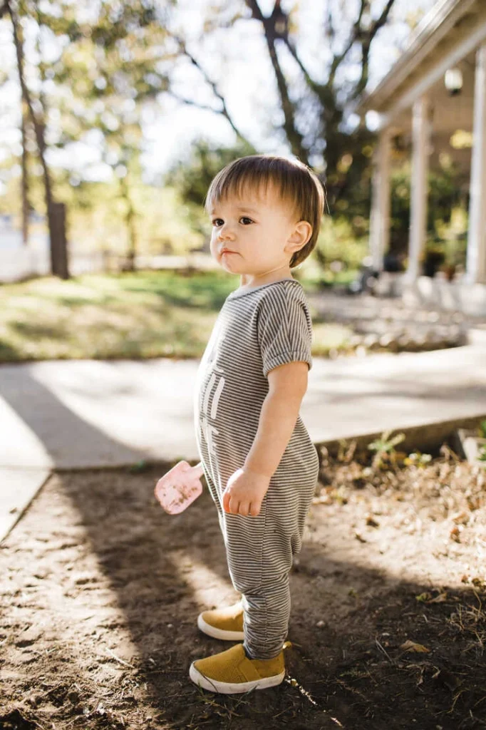 Cute baby boy in striped outfit stands outdoors, holding a pink toy, with sunlight filtering through trees.