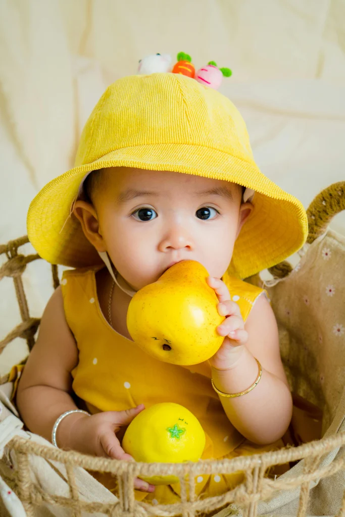 baby in a yellow hat holding a pear and lemon, sitting in a woven basket with a soft background