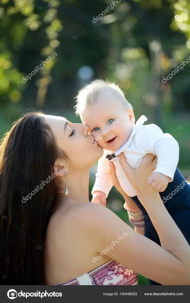 Mother kisses her smiling baby boy outdoors, surrounded by greenery and soft sunlight.