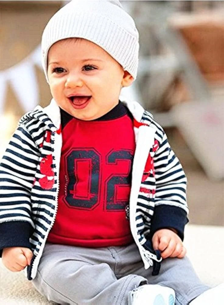 smiling baby in a striped hoodie and red shirt, wearing a white beanie, sitting happily outdoors