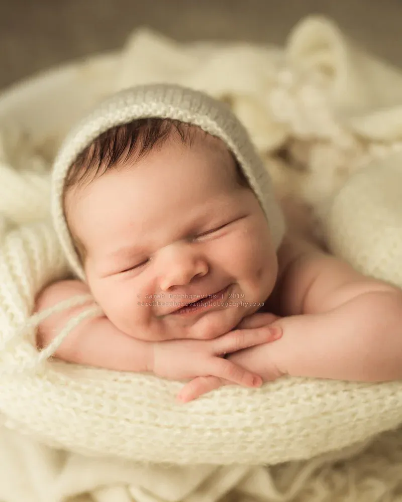 smiling baby with a knitted hat resting on a soft blanket, hands clasped under chin, exuding cuteness and warmth