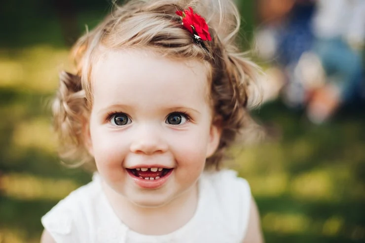 Smiling baby with curly hair and a red bow, enjoying a sunny day outdoors.