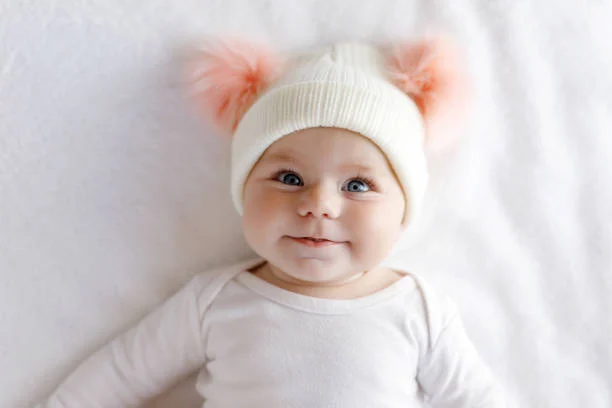 smiling baby wearing a white hat with pink pom-poms, lying on a soft white surface